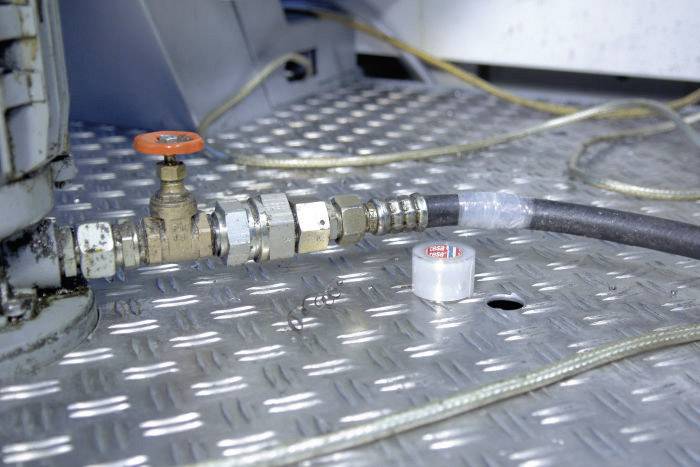 Ein Wasserhahn mit rotem Griff und angeschlossenem Schlauch auf einer geriffelten Metalloberfläche, im Hintergrund verlaufen Kabel.