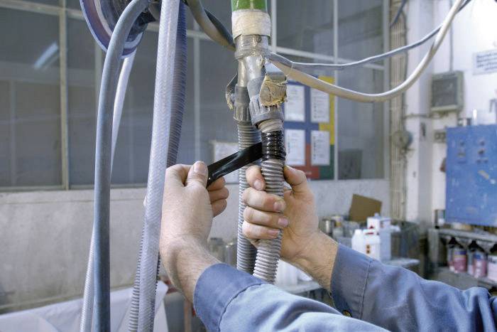 Eine Person repariert einen Schlauch in einer Werkstatt mit schwarzem Klebeband, um eine Verbindung zu sichern. Im Hintergrund sind Werkzeuge sichtbar.