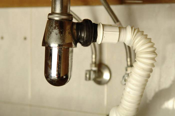 Ein verchromtes Siphon-Rohr unter einem Waschbecken, verbunden mit einem weißen, flexiblen Abflussrohr. Hintergrund: geflieste Wand.