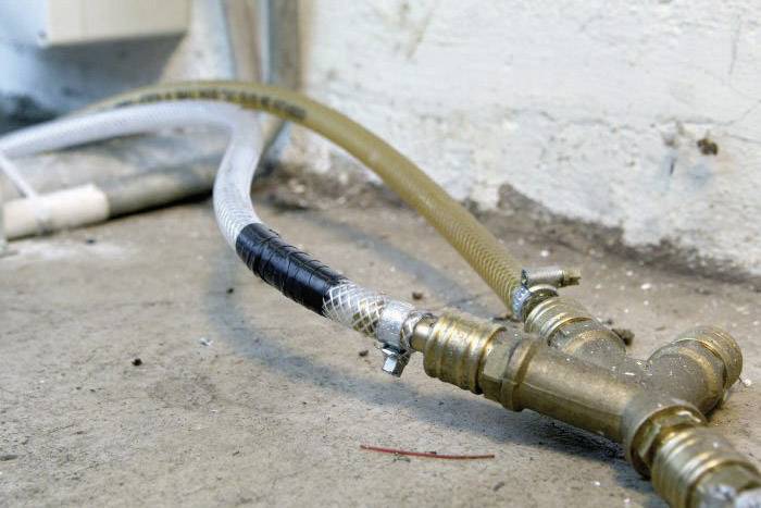 Ein Brauchwasserschlauch mit zwei Ventilen verbindet Rohrleitungen an einer Wand auf einem Betonboden.