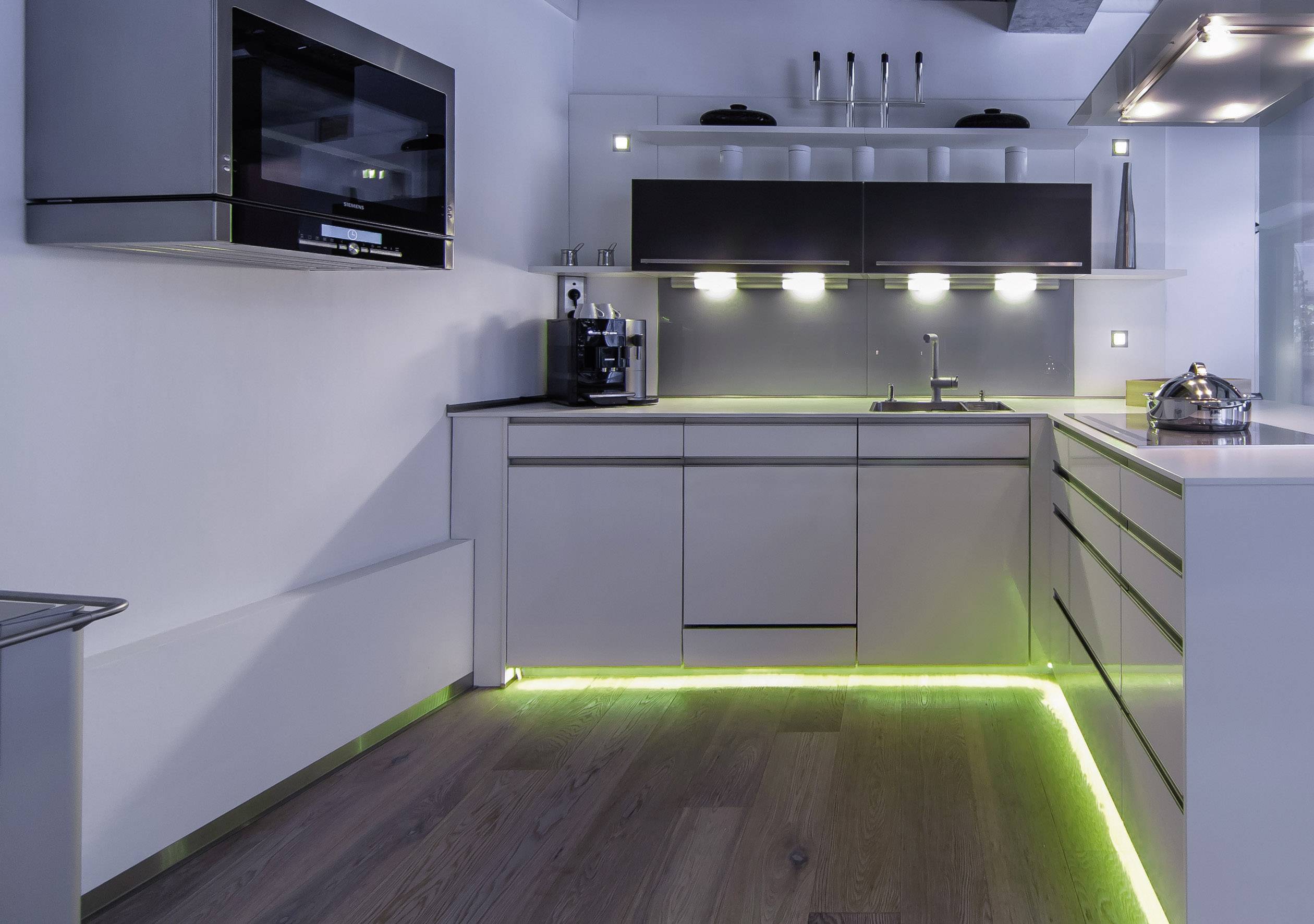 Moderne Küche in neutralen Tönen mit eingebauten Schränken und Geräten. Grüne LED-Beleuchtung am Boden erzeugt eine stilvolle Atmosphäre.