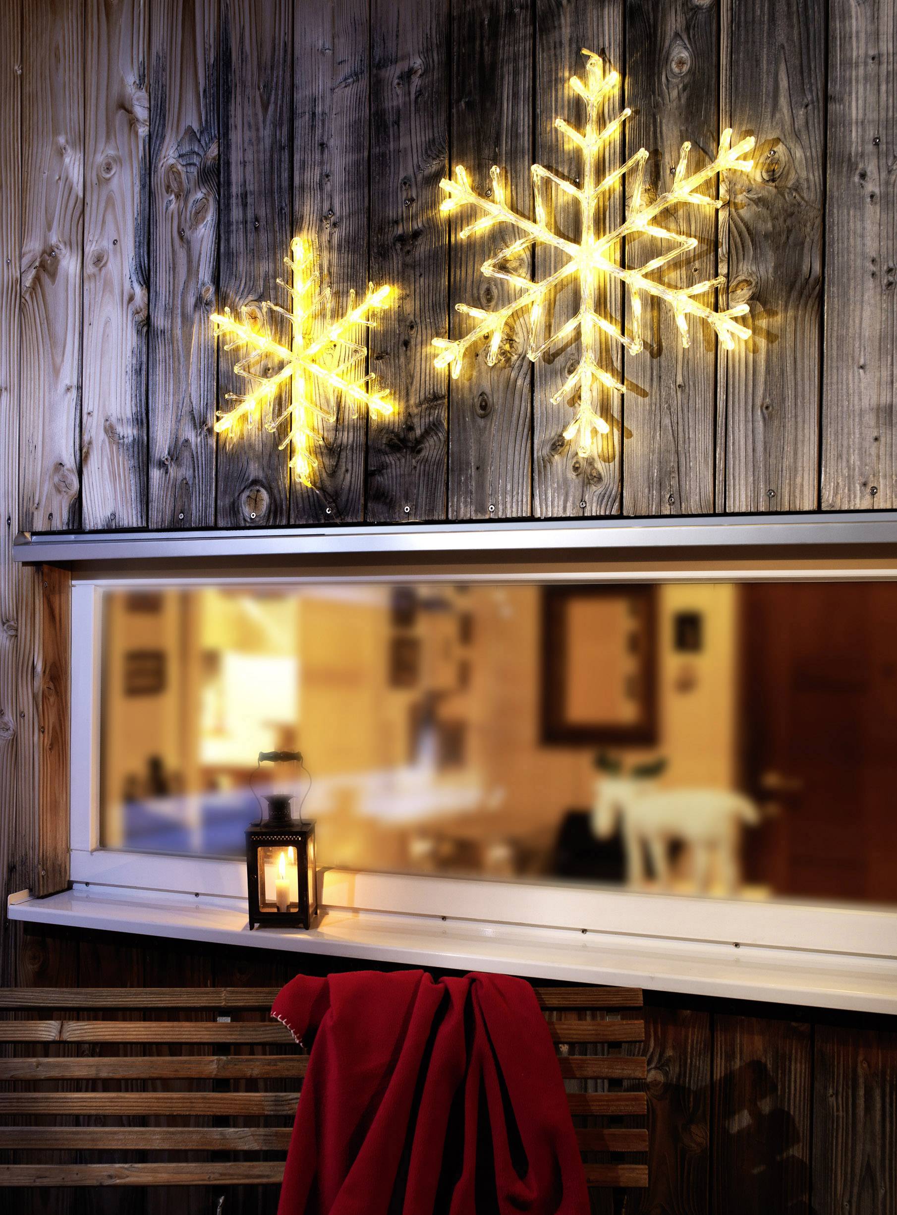 Lichterketten in Form von Schneeflocken leuchten an einer Holzwand über einem Fenster. Eine rote Decke liegt auf einer Bank.