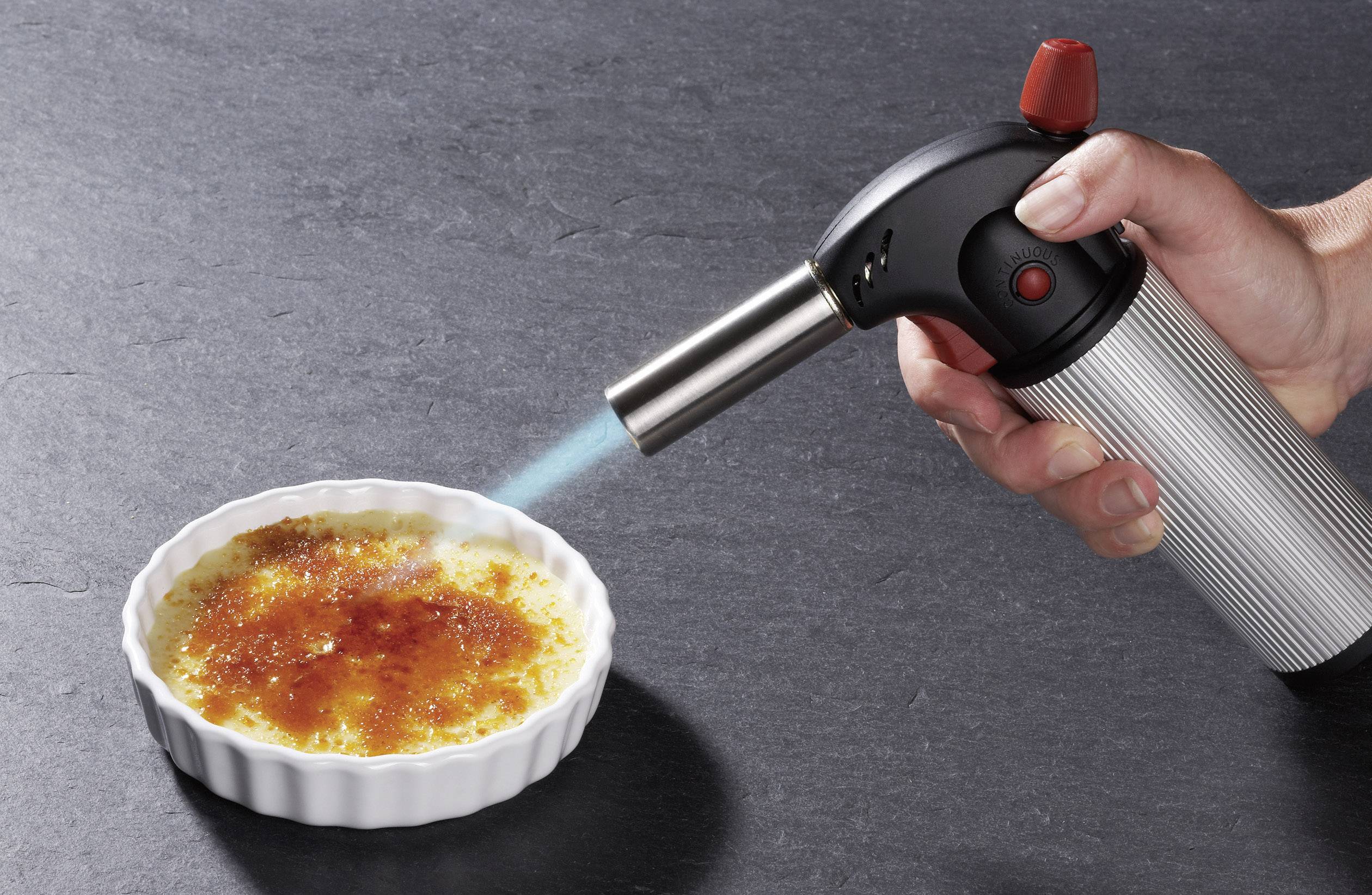 Hand hält Lötlampe über einer Crème Brûlée in einer weißen Schale, während die Oberfläche karamellisiert wird.