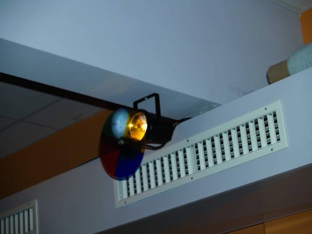Ein Scheinwerfer an der Decke projiziert Licht durch eine mehrfarbige Linse. Ein Lüftungsgitter ist daneben montiert.