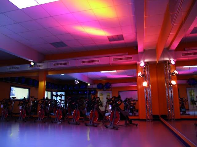 Fitnessstudio mit Neonlichtern. Der Raum ist mit mehreren stationären Fahrrädern ausgestattet, die vor einem Spiegel stehen.