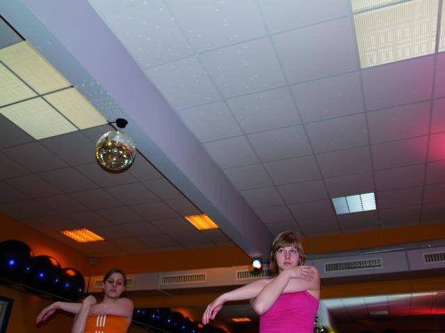 Zwei Personen in einem Fitnessstudio führen Tanzbewegungen aus, umgeben von Spiegeln, mit einem Discoball an der Decke.