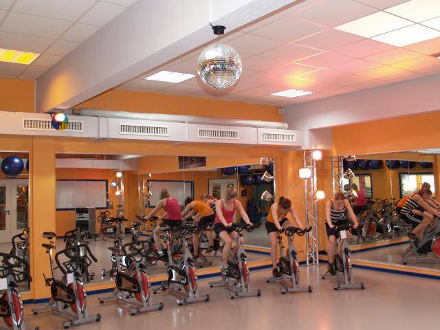 Personen trainieren auf stationären Fahrrädern in einem Fitnessstudio mit Spiegelwänden und Discolicht.
