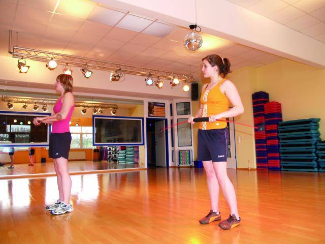 Zwei Personen springen in einem Fitnessstudio mit einem Springseil. Im Hintergrund spiegeln Spiegel den Raum wider, bunte Matten sind stapelweise gestapelt.