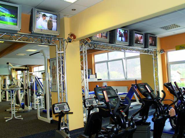 Fitnessstudio mit Laufbändern und Fahrradergometern. An der Wand hängen mehrere Fernseher, auf denen unterschiedliche Programme laufen.