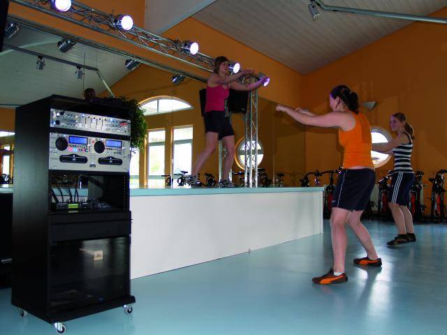 Eine Aerobic-Trainerin leitet eine Gruppe von drei Personen bei einem Fitness-Workout in einem Studio mit Spiegelwand und Lautsprecherturm.