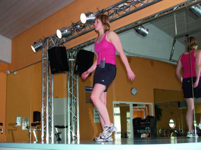 Eine Frau in pinkem Top und schwarzen Shorts macht Aerobic auf einer Bühne in einem Fitnessstudio, umgeben von Scheinwerfern und Spiegeln.