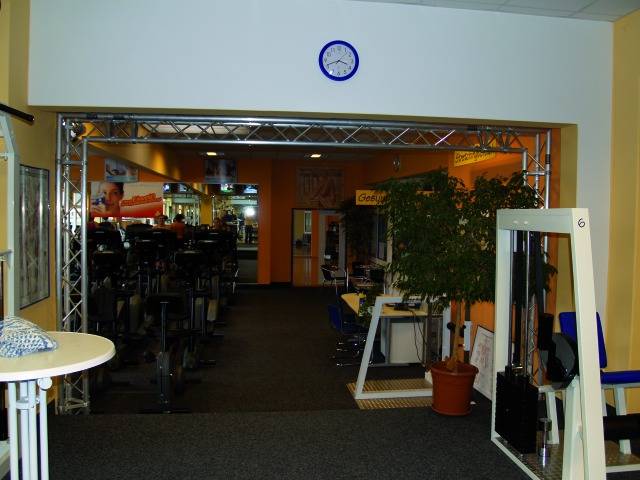 Innenraum eines Fitnessstudios mit mehreren Fitnessgeräten, gelben Wänden, einer grünen Pflanze und einer Wanduhr über einem Eingang.