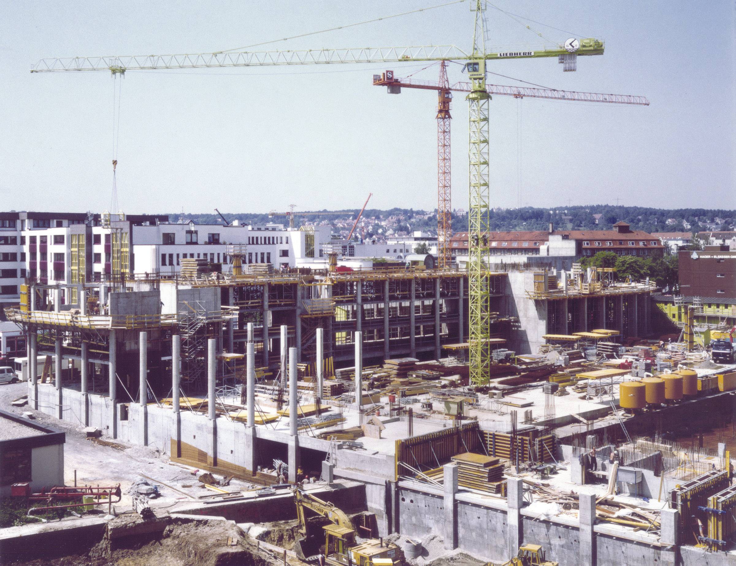 Eine Baustelle mit großen Kränen und einem im Bau befindlichen mehrstöckigen Gebäude. Arbeiter und Materialien sind sichtbar.