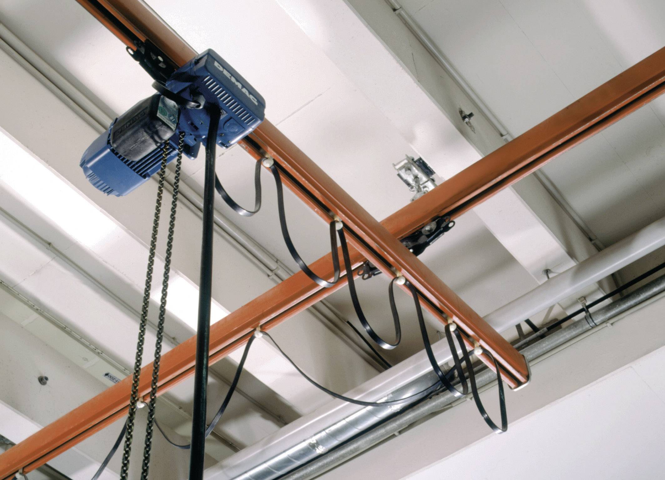 Eine blaue elektrische Kettenzugmaschine ist an einer orangenen Schiene an der Decke befestigt und wird durch schwarze Kabel verbunden.