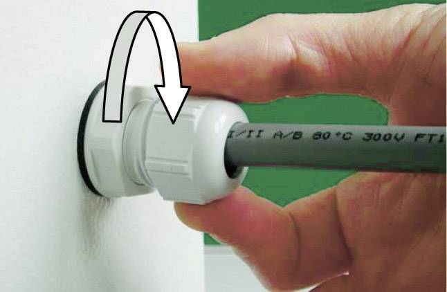 Hand zieht Kabel durch weiße Verschraubung in der Wand. Pfeil zeigt Drehrichtung zum Festziehen. Kunststoffklemmung sichert das Kabel.