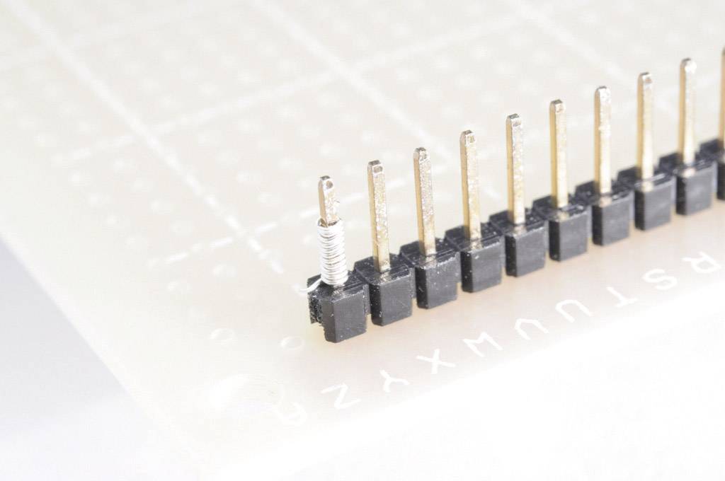 Eine schwarze, einreihige Stiftleiste ist auf einer Steckplatine montiert, links mit einer gedrehten Drähtenwicklung an einem Stift.