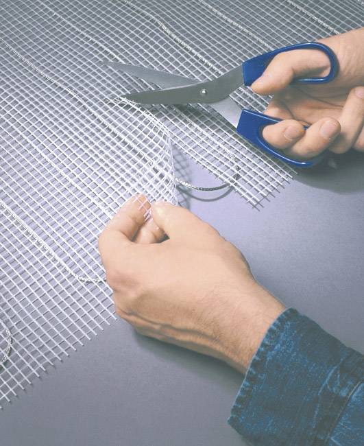 Eine Person schneidet ein netzartiges Material mit einer blauen Schere. Die Hände sind im Fokus und zeigen den Schneidevorgang.