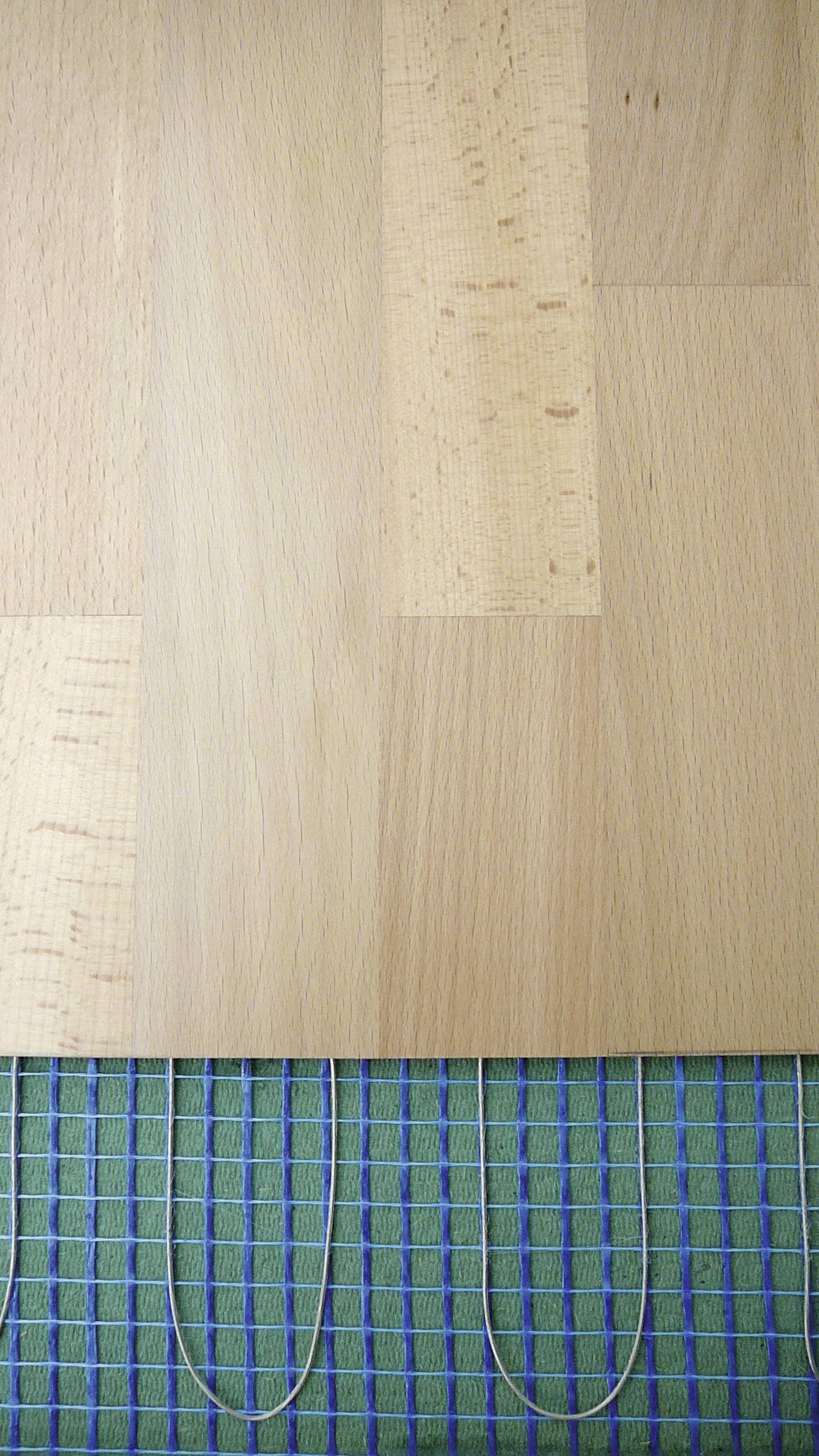 Holzfußboden mit sichtbarer blauer Heizmatte darunter, die Heizdrähte auf grünem Untergrund zeigt.