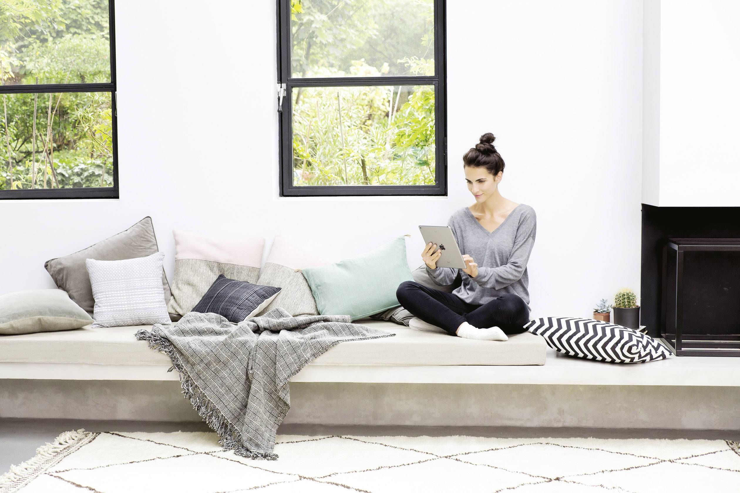 Eine Frau sitzt gemütlich auf einem großen modernen Sofa mit vielen Kissen und liest auf ihrem Tablet. Im Hintergrund sind Fenster.