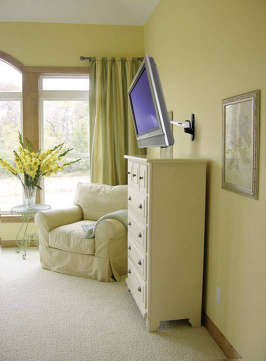 Ein gelber Raum mit einem Sessel und einem hohen Schrank, auf dem ein Flachbildfernseher befestigt ist. Ein Blumenarrangement steht daneben.