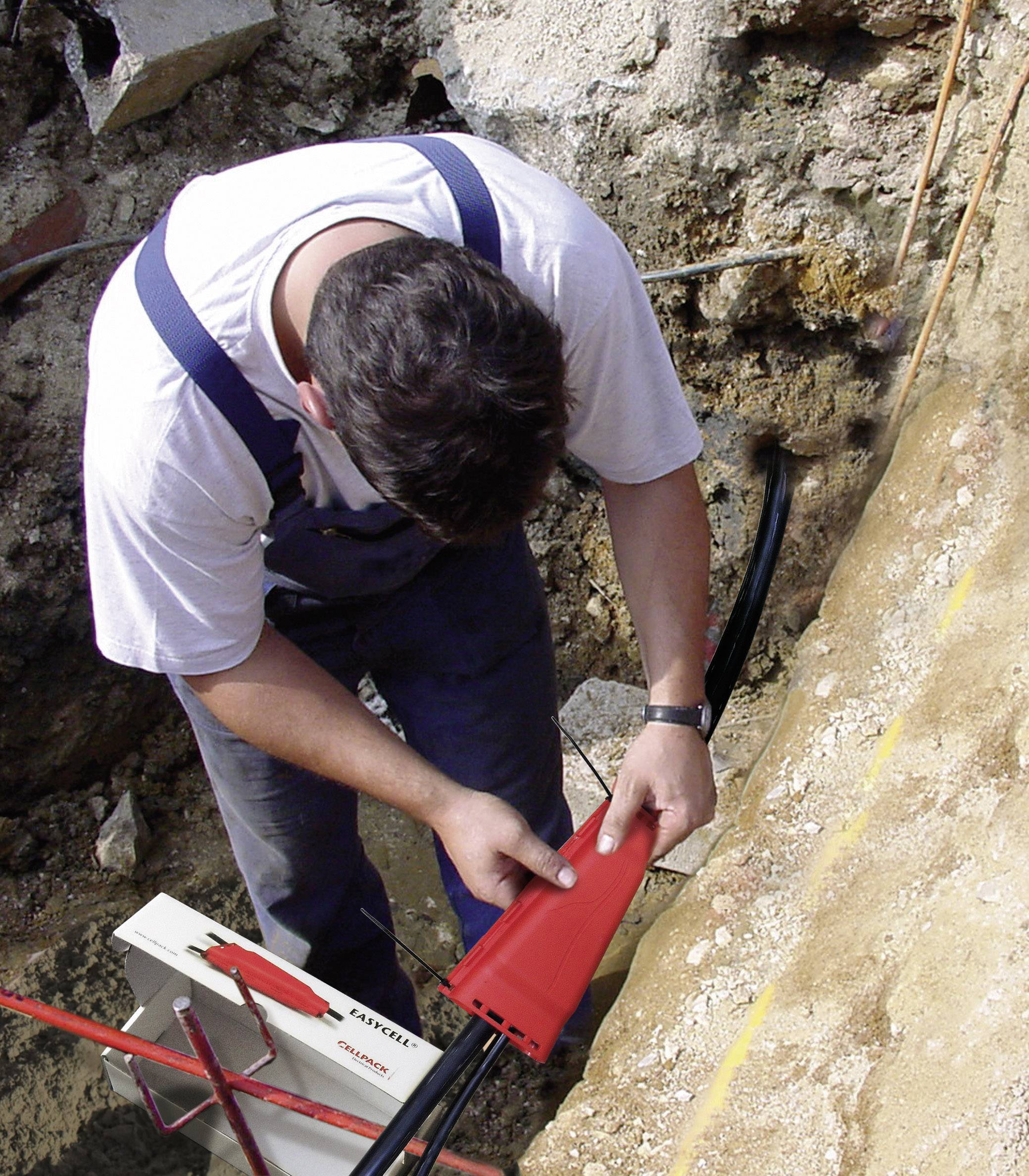 Ein Arbeiter in Arbeitskleidung verlegt Kabel im Erdboden und prüft ein rotes Schutzrohr. Umgebungsbedingungen deuten auf Bauarbeiten hin.