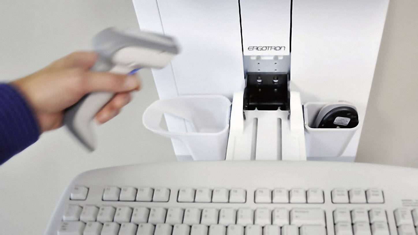 Eine Hand hält einen Barcodescanner über einer weißen Tastatur auf einem Schreibtisch. Rechts befinden sich Ablagen für Bürobedarf.