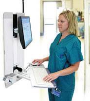 Eine Person in medizinischer Kleidung arbeitet an einem Wandcomputer in einem Krankenhausflur. Sie tippt auf einer Tastatur.