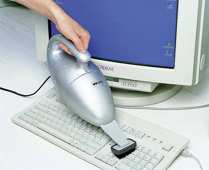 Eine Hand reinigt eine Computertastatur mit einem tragbaren Staubsauger vor einem Monitor auf einem Schreibtisch.