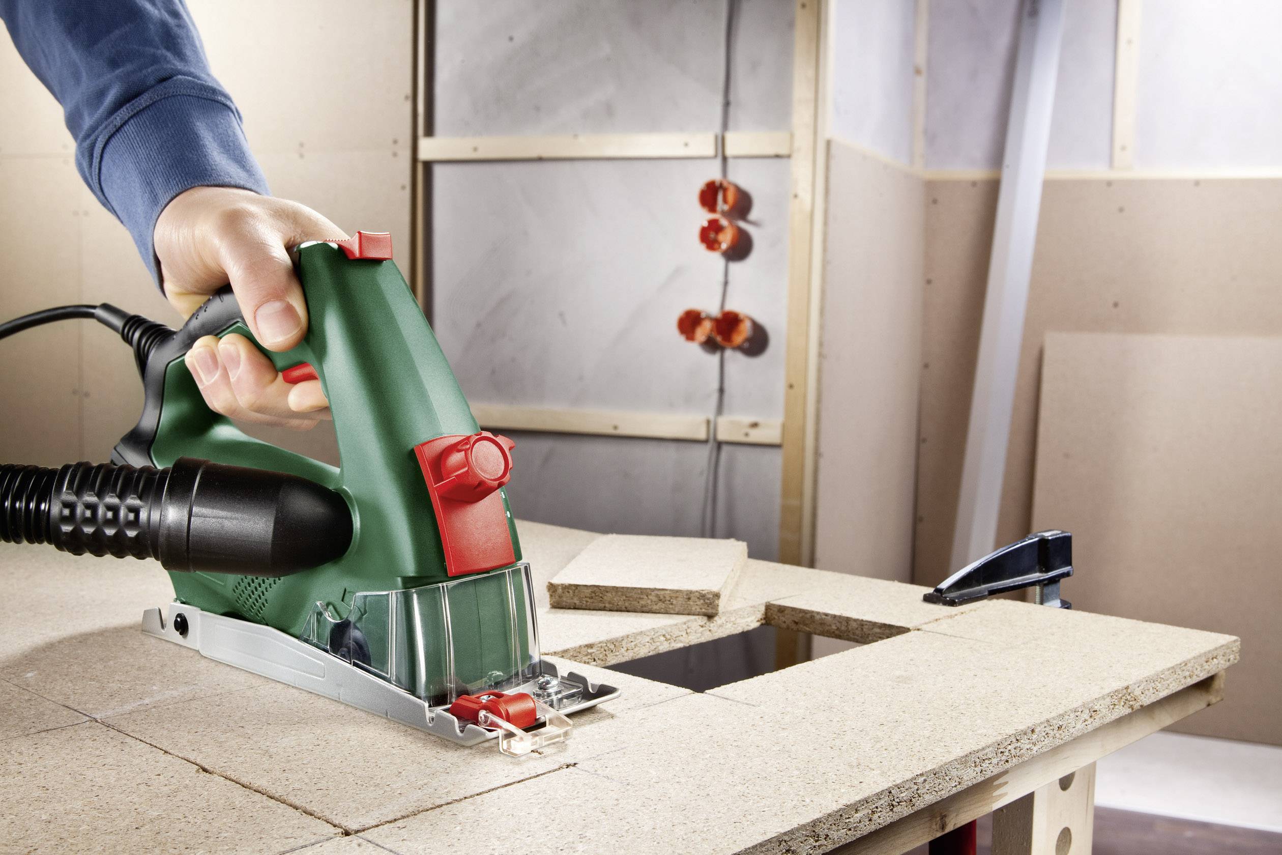 Eine Person bedient eine grüne elektrische Säge und schneidet ein Holzbrett auf einem Arbeitstisch in einer Werkstattumgebung.