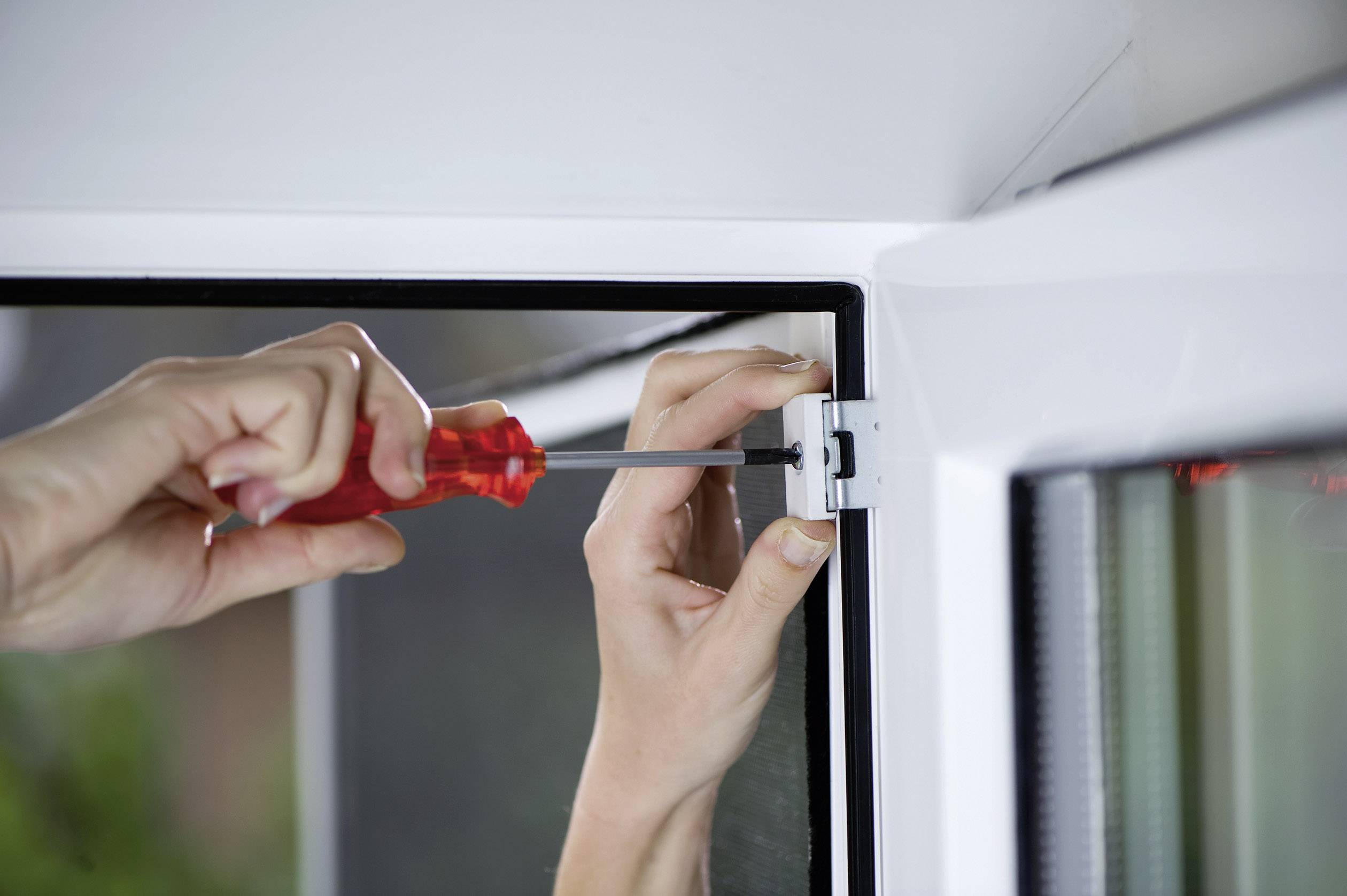 Hände reparieren ein Fenster mit einem Schraubenzieher, während eine Person das Scharnier anzieht, um die Stabilität zu gewährleisten.