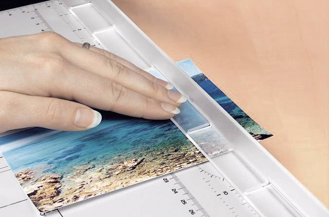 Eine Hand schneidet ein Foto von einer Küstenlandschaft mit einem Papierschneider. Ein Lineal zeigt Zentimeter und Inch.