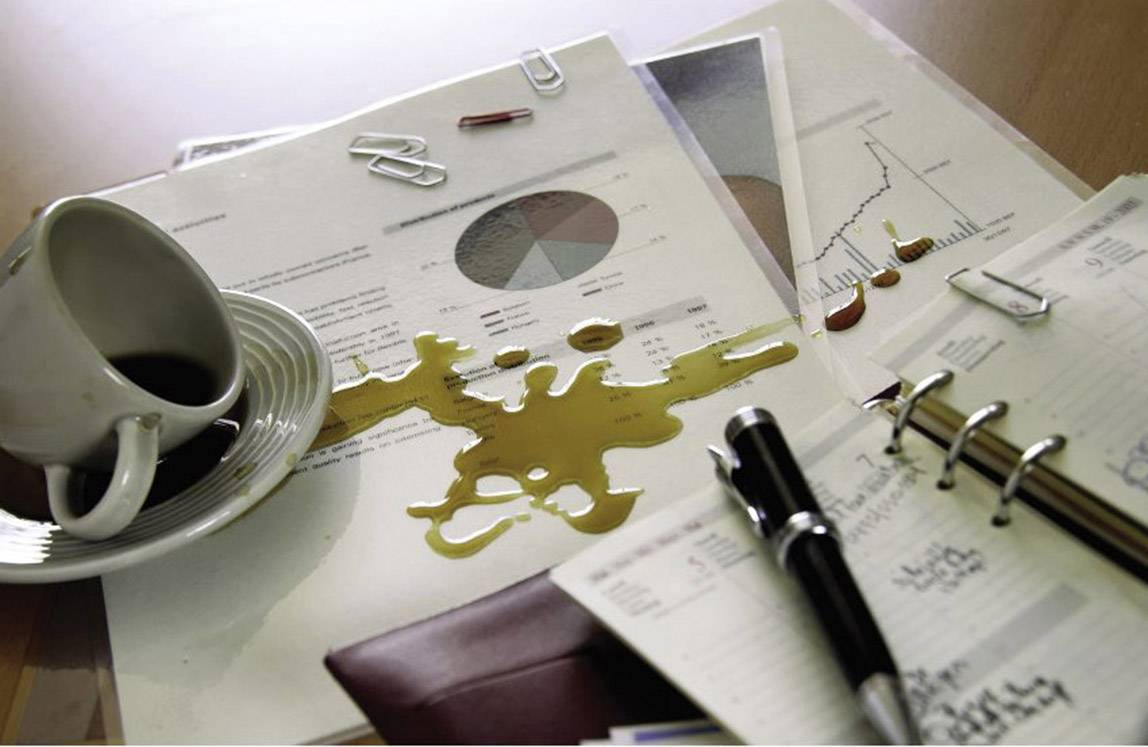 Eine umgestoßene Tasse Kaffee hat auf einem Schreibtisch Papiere mit Diagrammen und Grafiken verschüttet. Ein Notizbuch liegt daneben.