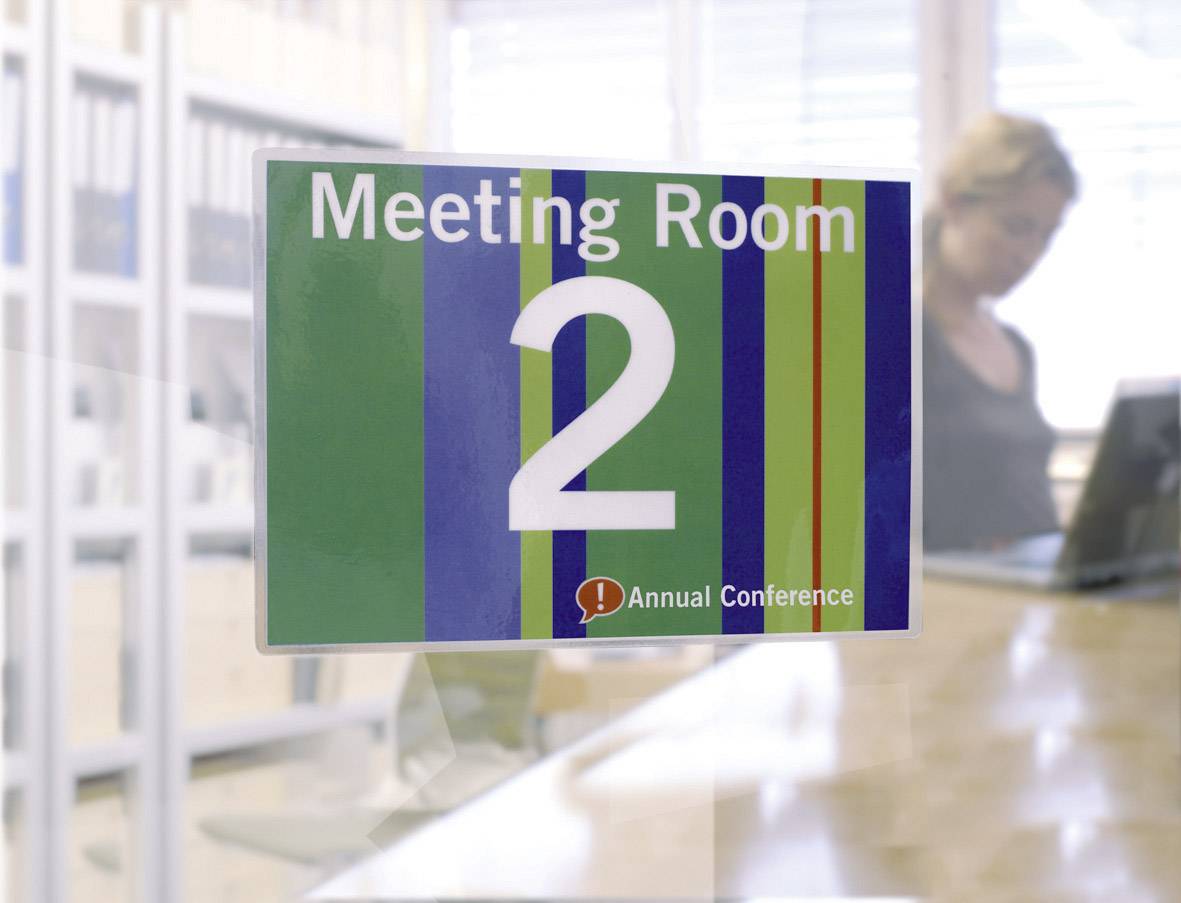 Ein Schild mit der Aufschrift 'Meeting Room 2' und 'Annual Conference' auf gestreiftem Hintergrund. Im Hintergrund ist eine unscharfe Person.