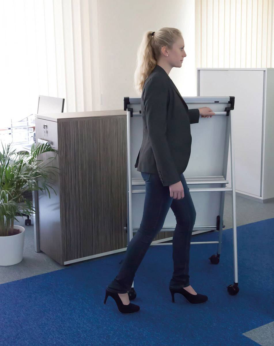 Eine Frau in formeller Kleidung zieht ein weißes Flipchart auf Rollen durch ein Büro mit blauen Teppichboden und Pflanzen im Hintergrund.