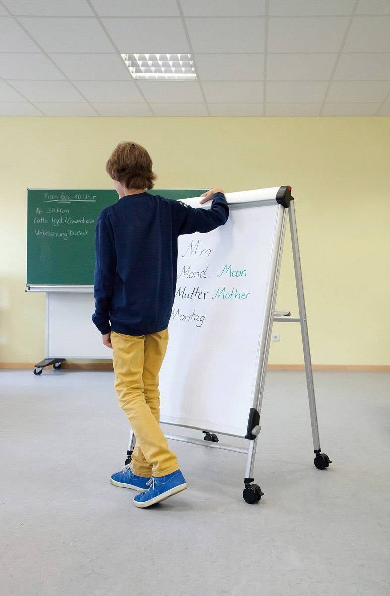 Ein Junge steht vor einem Whiteboard mit dem Buchstaben 'M'. Im Hintergrund ist eine Tafel mit Aufschrift und Kreidezeichnungen.
