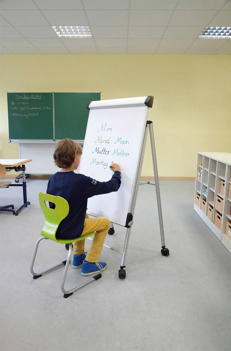 Ein Junge sitzt auf einem Stuhl in einem Klassenzimmer und schreibt Wörter, die mit 'M' beginnen, auf ein Flipchart.
