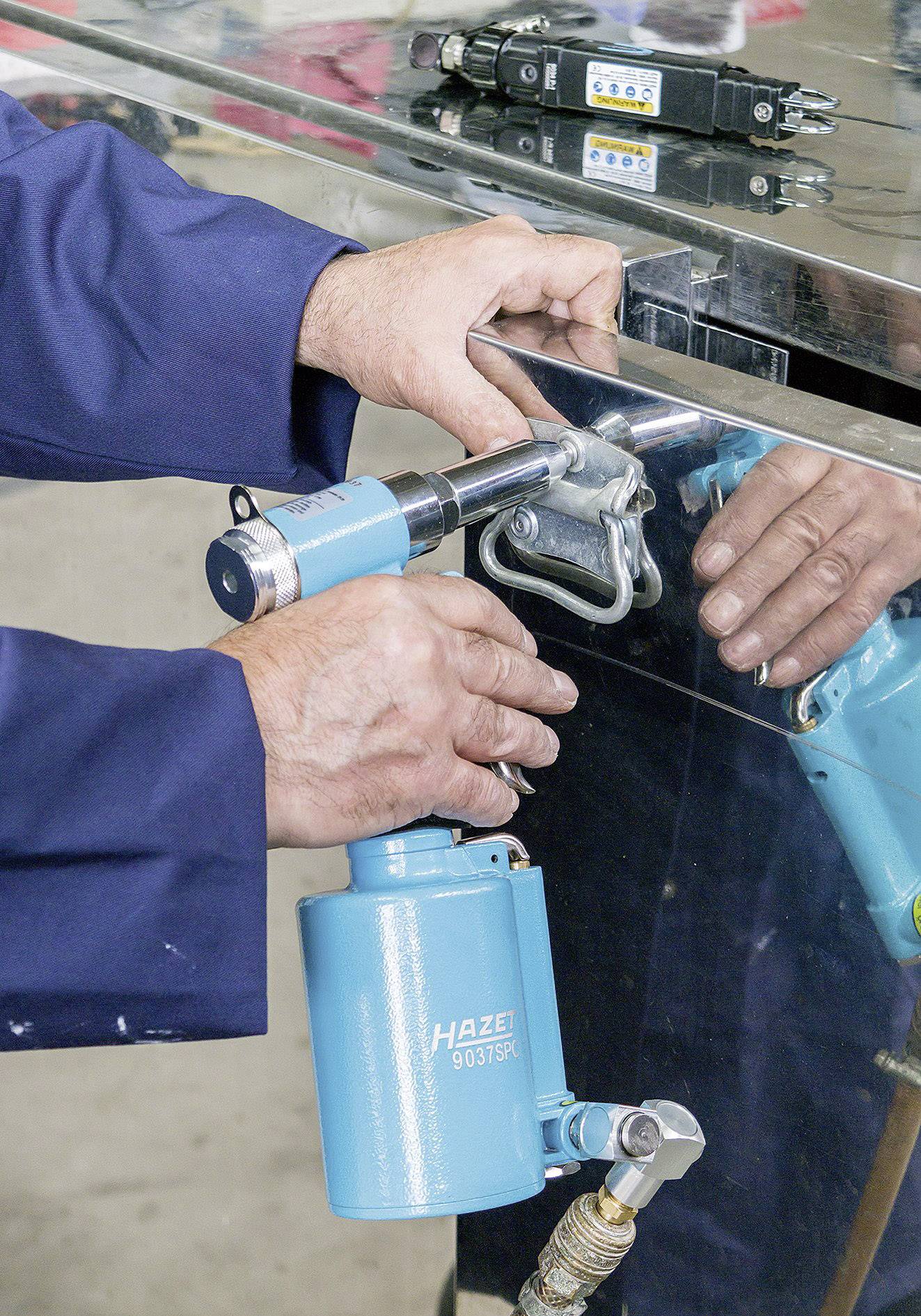 Ein Arbeiter in Arbeitskleidung benutzt ein blaues Werkzeug, um eine Klemme an einer Metalloberfläche zu befestigen. Reflektionen sichtbar.