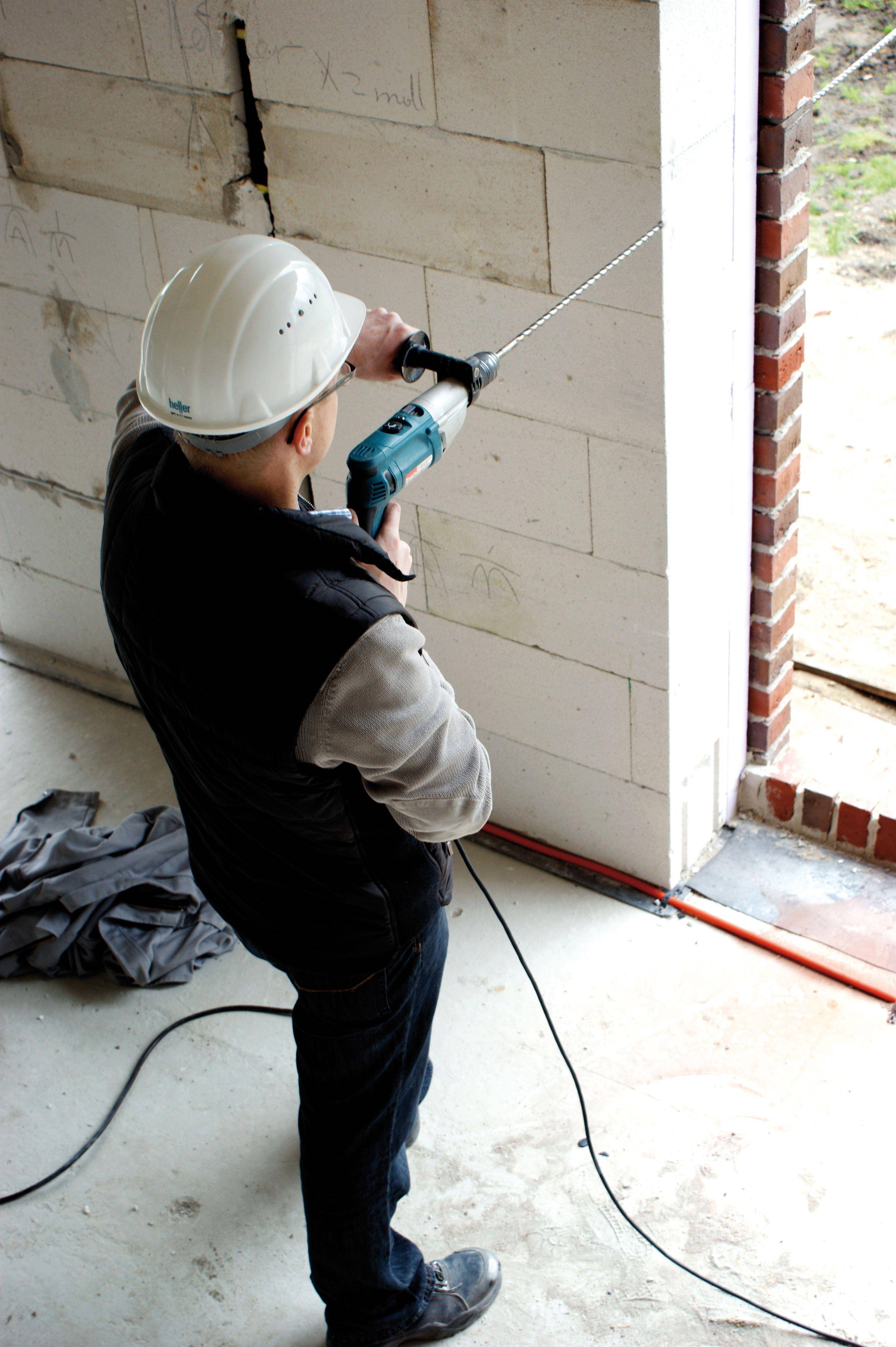 Arbeiter mit Schutzhelm nutzt Bohrer an Mauer in Baustelle.