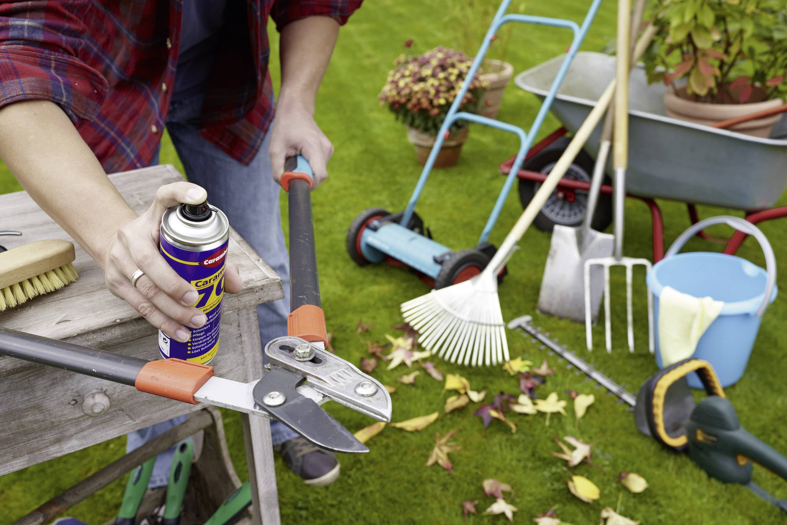 Eine Person pflegt Gartenwerkzeuge mit einem Schmiermittel. Im Hintergrund sind verschiedene Gartengeräte und Herbstlaub zu sehen.