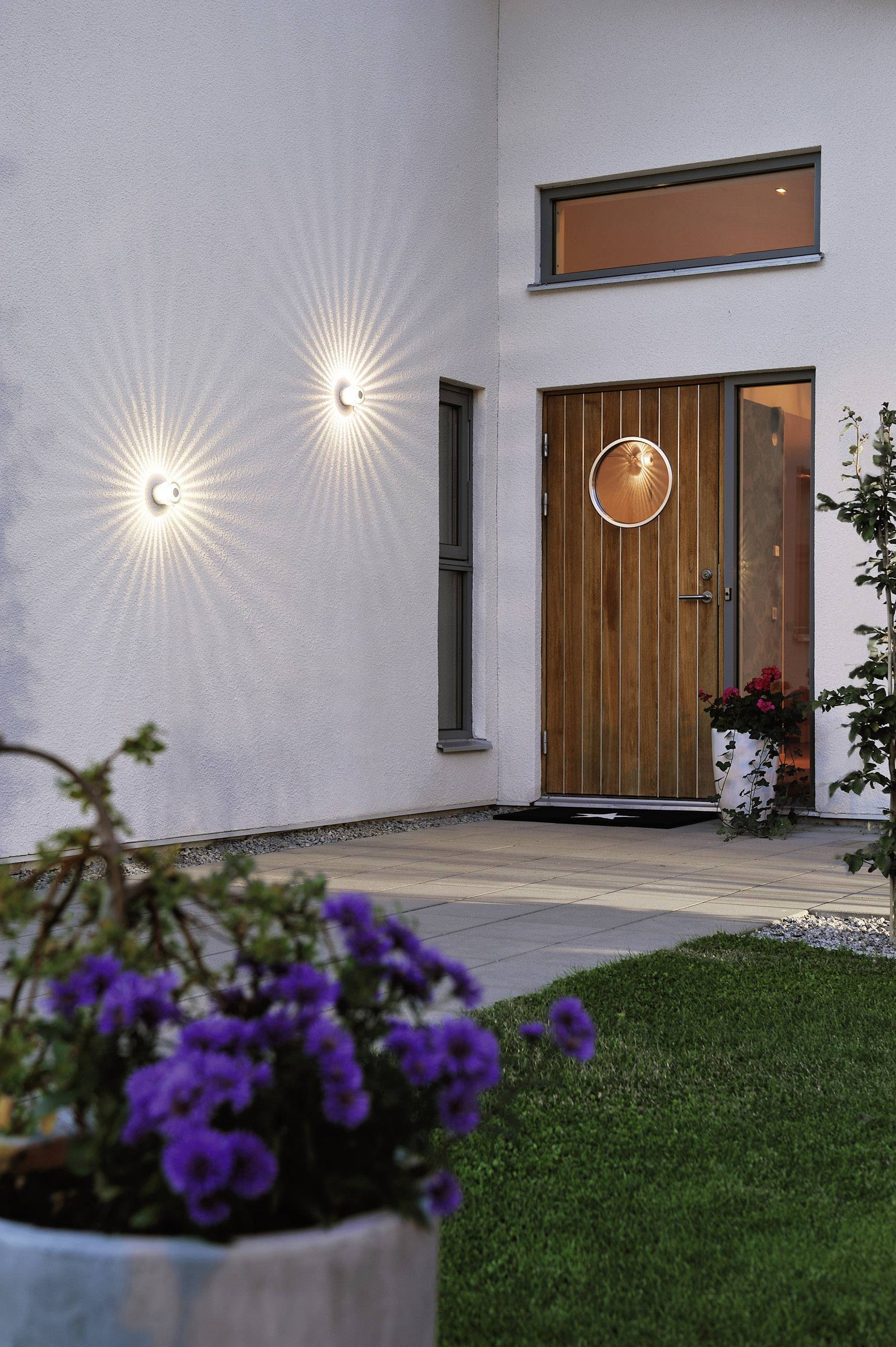 Garten mit beleuchteter Hausfassade, Holztür, und Blumen im Vordergrund, vermittelt eine einladende Abendstimmung.
