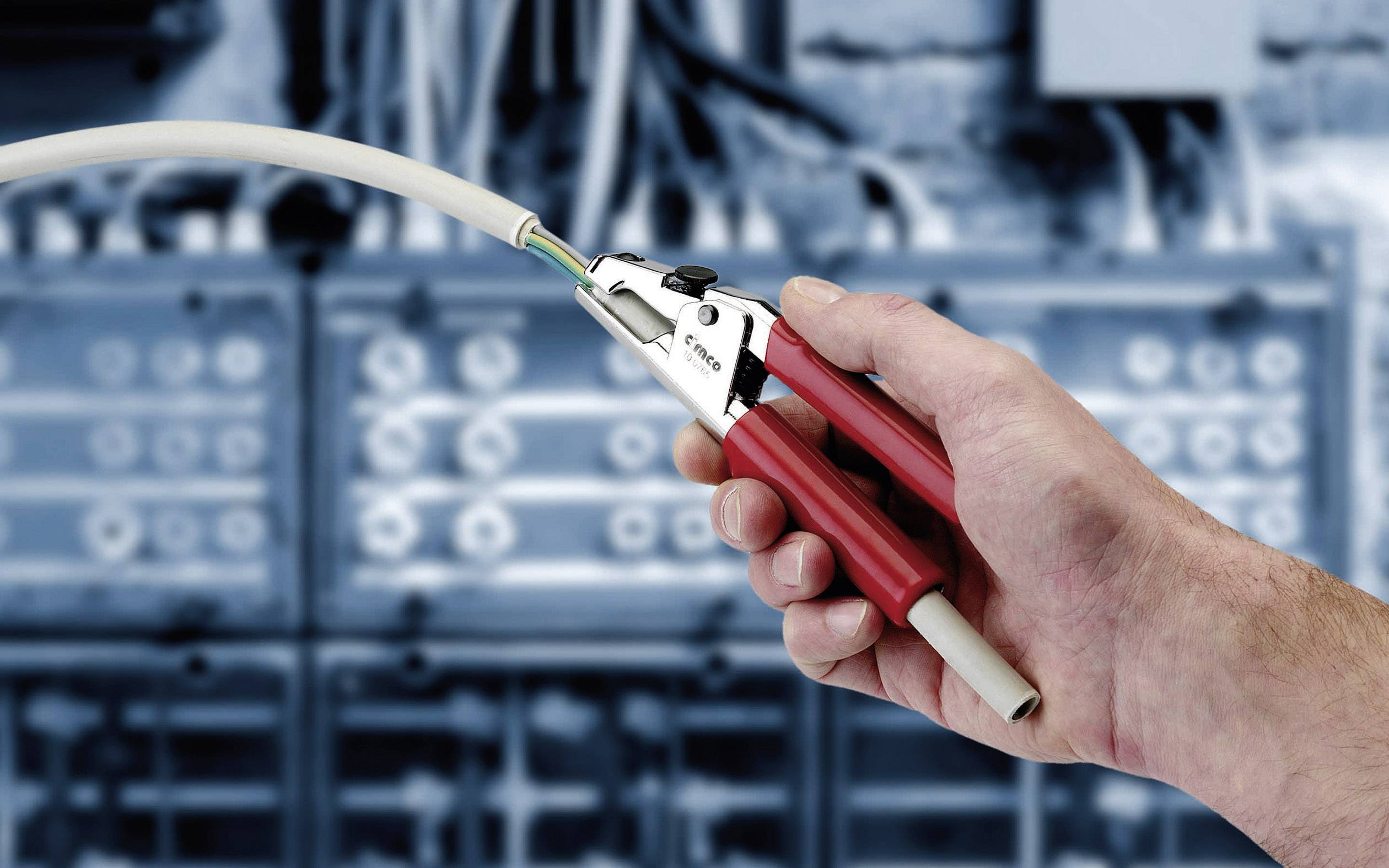 Hand hält ein Werkzeug zum Abisolieren von Kabeln vor einem unscharfen Hintergrund mit elektrischen Schaltkästen.