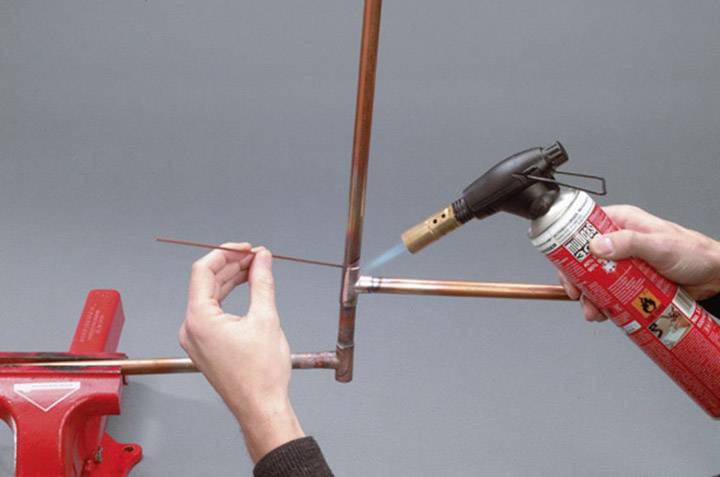 Eine Person lötet Kupferrohre mit einem Gasbrenner. Ein weiteres Rohr wird mit der linken Hand gehalten. Fokus auf handwerkliche Tätigkeit.