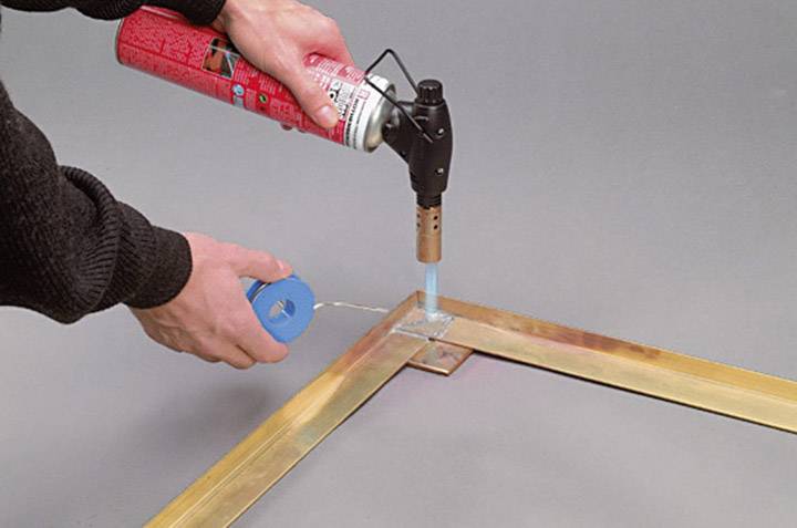 Zwei Hände, die eine Lötlampe und Lötzinn benutzen, um zwei Metallstücke an einer Ecke zu verbinden.