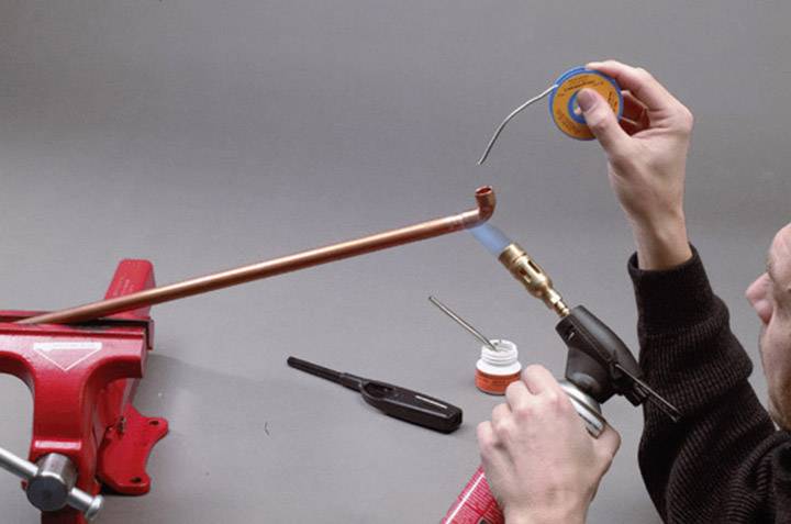 Eine Person lötet ein Kupferrohr, das in einem roten Schraubstock fixiert ist. Sie hält einen Brenner in der rechten Hand und Lot in der linken.