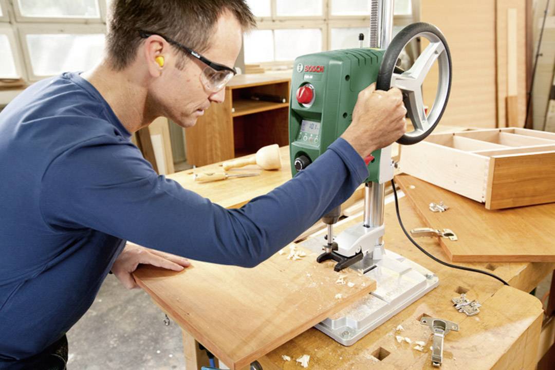 Ein Mann in Schutzbrille arbeitet an einer Tischbohrmaschine in einer Werkstatt, konzentriert auf das Bohren eines Holzstücks.