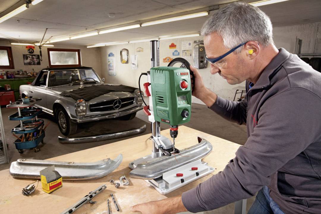 Ein Mann arbeitet in einer Werkstatt an einem Bohrständer, während im Hintergrund ein klassisches Auto steht.