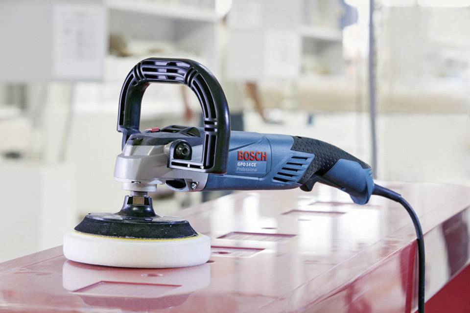 Elektroschleifer in Werkstattumgebung auf einem Metalltisch. Das Gerät ist blau mit schwarzem Griff und weißem Schleifpad.