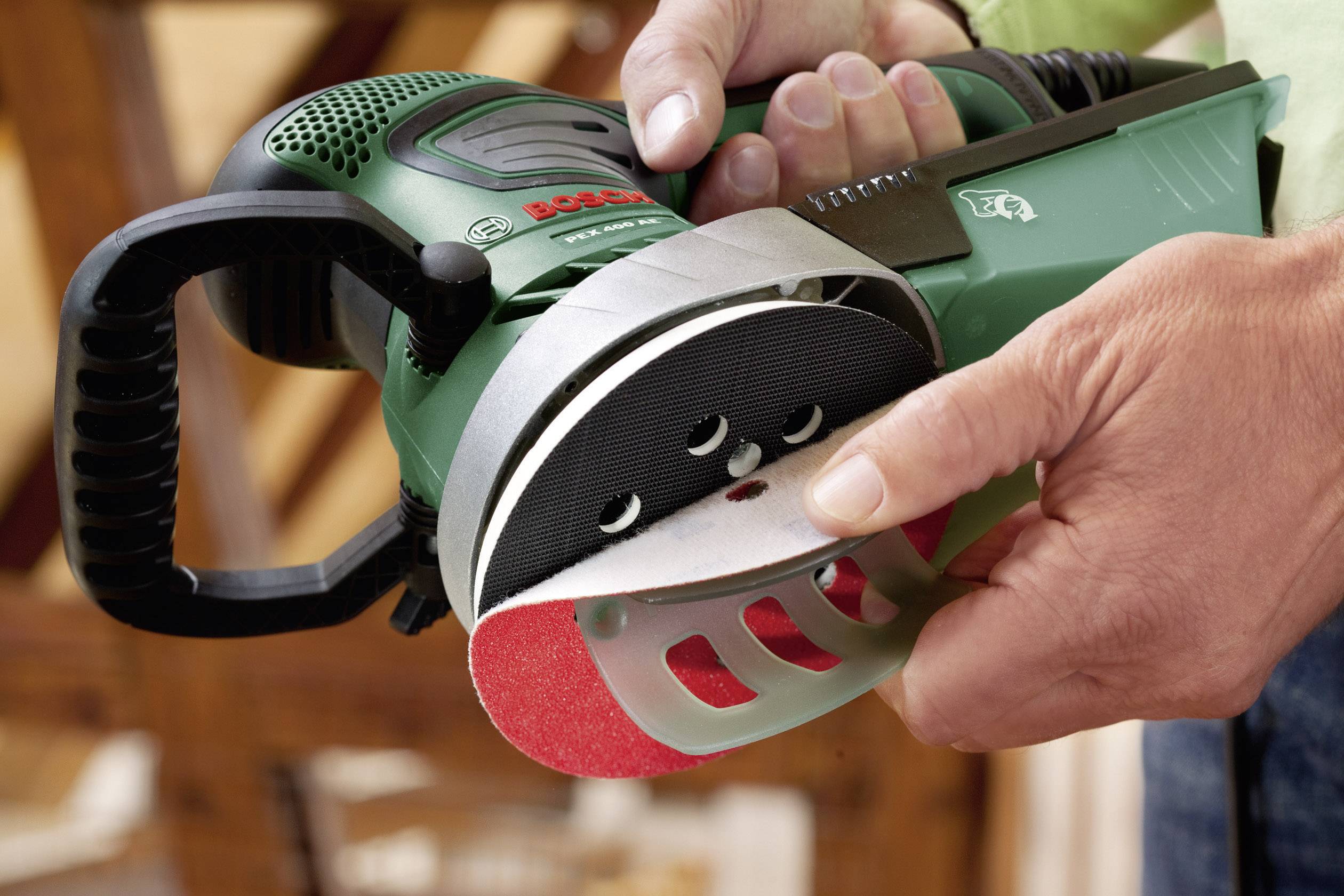Eine Person wechselt das Schleifpapier an einem grünen Bosch-Schleifgerät. Hände halten das Gerät und das rote Schleifpapier.