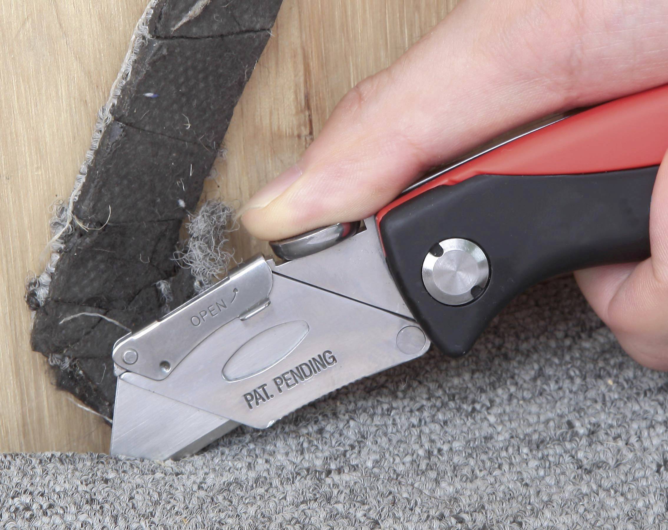 Eine Hand verwendet ein Teppichmesser, um einen Teppich an einer Kante zu schneiden. Die Klinge ist halb ausgefahren.