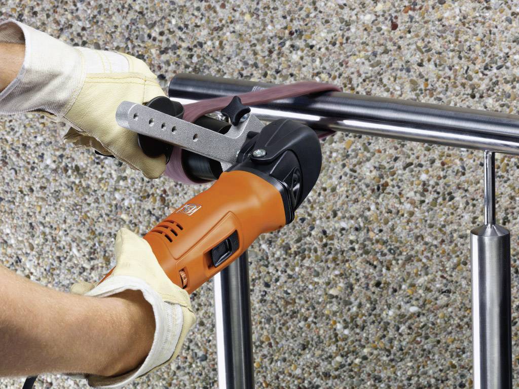 Eine Person schleift ein Metallgeländer mit einem Winkelschleifer, tragend Handschuhe für Sicherheit. Im Hintergrund ist eine Steinmauer sichtbar.